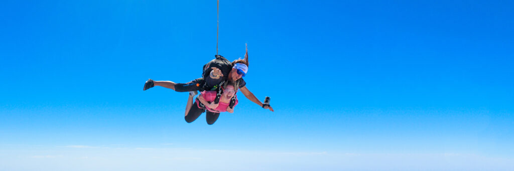 A tandem skydive pair is free-falling against a bright blue sky. The instructor is wearing a black suit, while the person in front is in a pink outfit. Both are facing downwards, with arms outstretched, capturing the thrill of the descent.