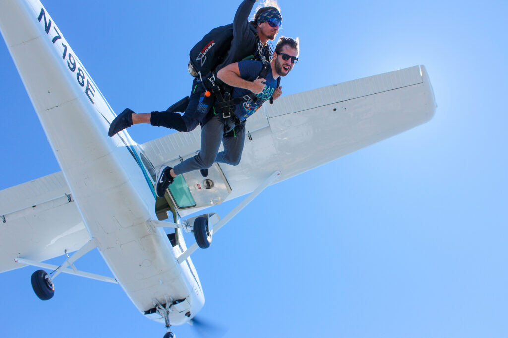 Two people tandem skydiving from a small airplane against a clear blue sky. The instructor is behind, and both are equipped with parachutes, experiencing free fall after jumping from the plane.