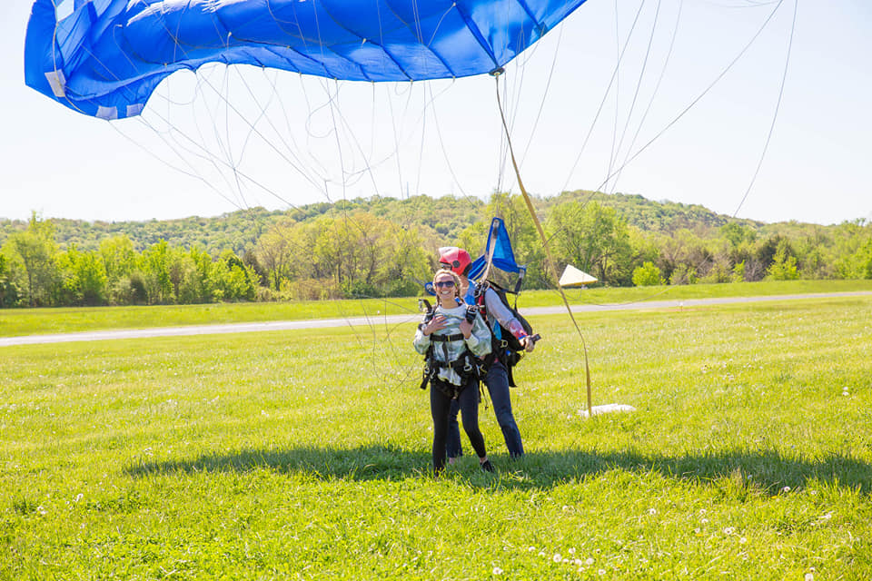 Two people land with a blue parachute on a grassy field. The tandem skydivers are smiling and wearing harnesses and helmets. Trees and a hill are in the background under a clear, sunny sky.