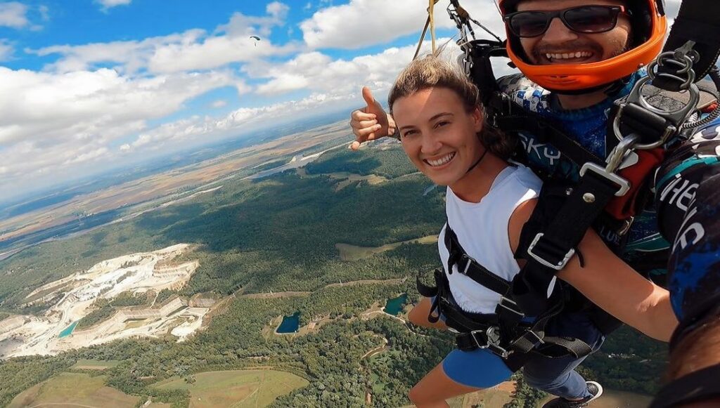 Two people tandem skydiving, smiling at the camera, with one giving a thumbs-up. They are descending over a landscape with forests, fields, and a quarry visible below. The sky is partly cloudy.