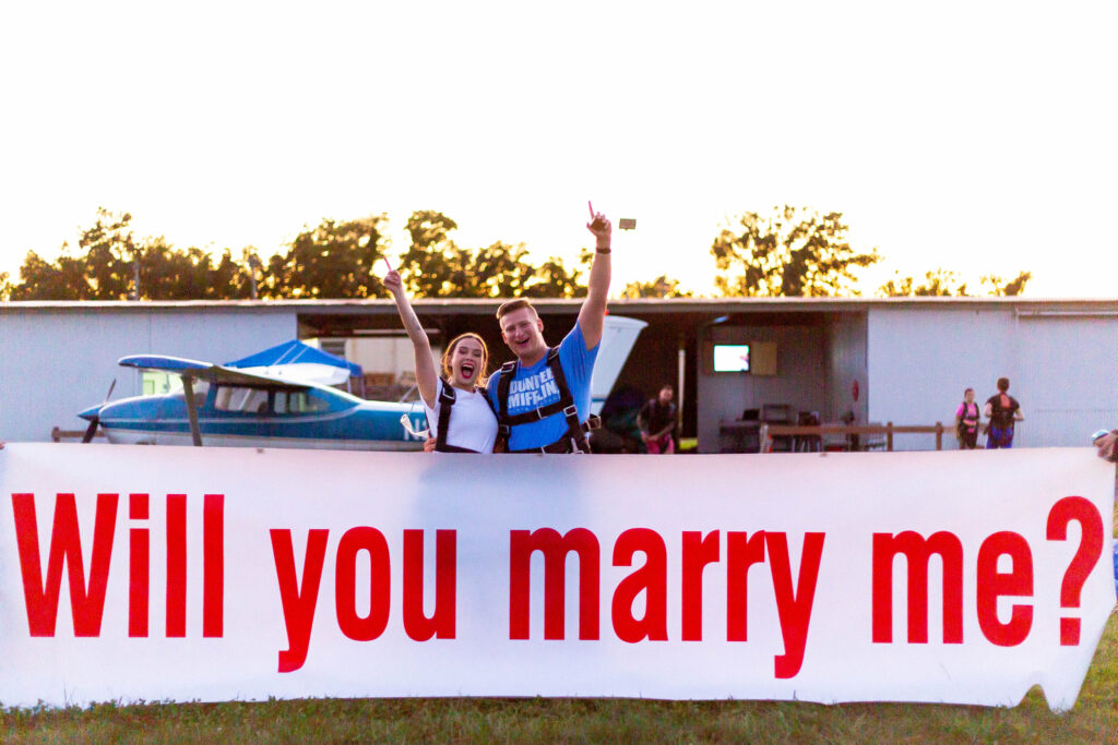 A couple stands joyfully in front of a small plane, holding a large banner that reads, "Will you marry me?" The sun is setting, casting a warm glow.