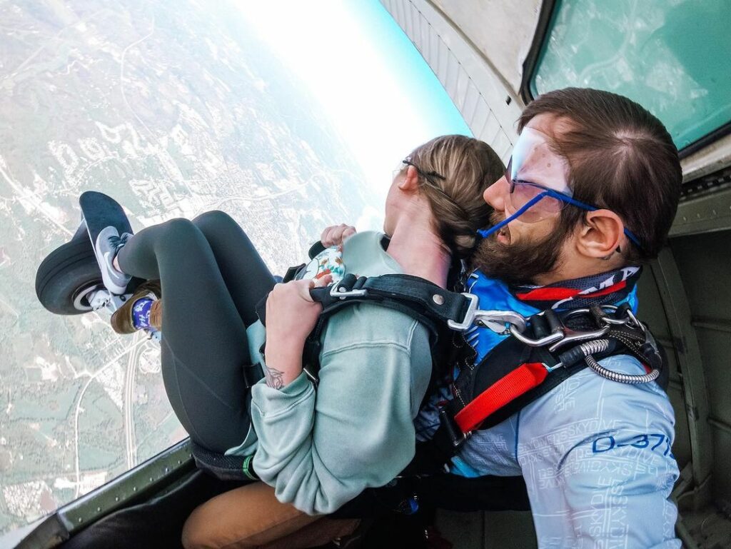 Two people are tandem skydiving, sitting at the open side of a plane. They wear harnesses and goggles. The landscape with fields and roads is visible below them. Both seem ready to jump out of the aircraft.