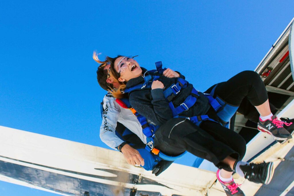 Two people tandem skydiving, mid-air after jumping from a plane. The person in front wears glasses and a blue harness, smiling with excitement. The instructor is behind, guiding the descent. The sky is clear and bright blue.