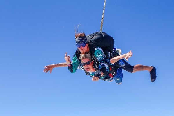 Two people are tandem skydiving against a clear blue sky. Both are wearing harnesses and casual clothes. The instructor is behind, while the other person is in front, with both expressing excitement.